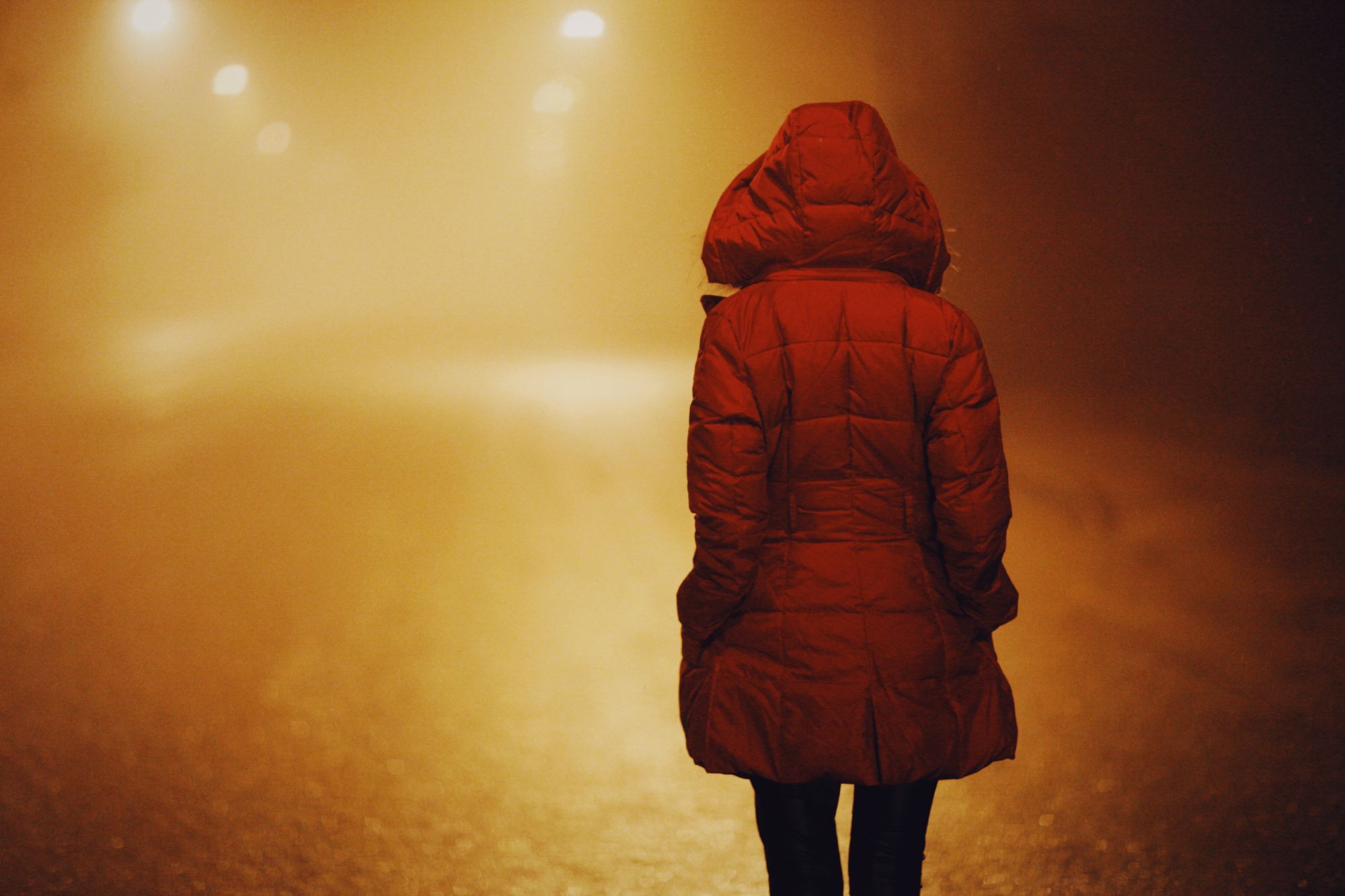 Person in a hooded puffer jacket walking on a foggy road at night under streetlights